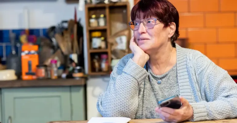 Mulher madura pensativa com o celular e papéis na mesa