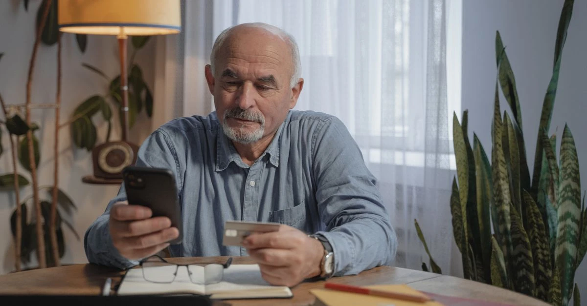 Aposentado sentado à mesa consultando o celular com um cartão na mão