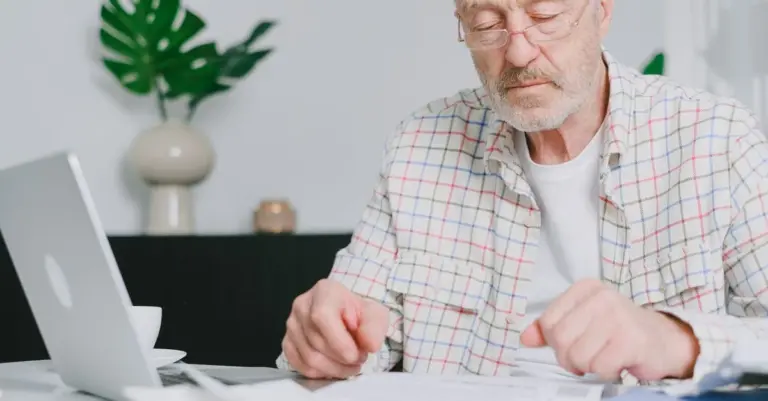 Homem idoso de óculos analisando documentos com o notebook