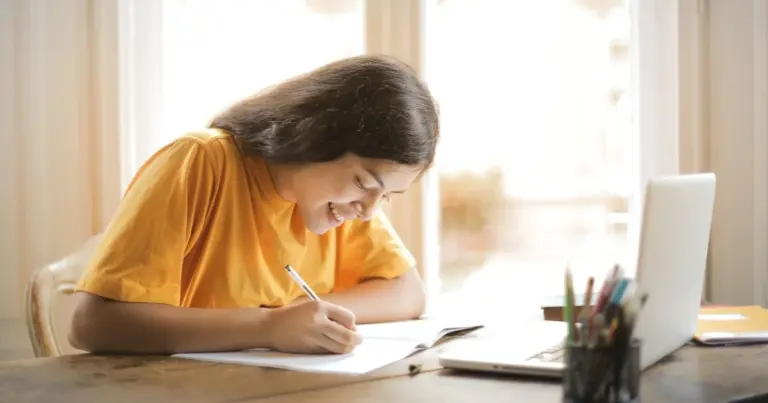 Jovem sorridente a escrever num caderno com laptop numa mesa de madeira