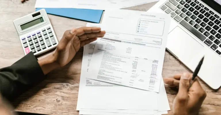 Pessoa segura documentos financeiros junto a calculadora e laptop numa secretária