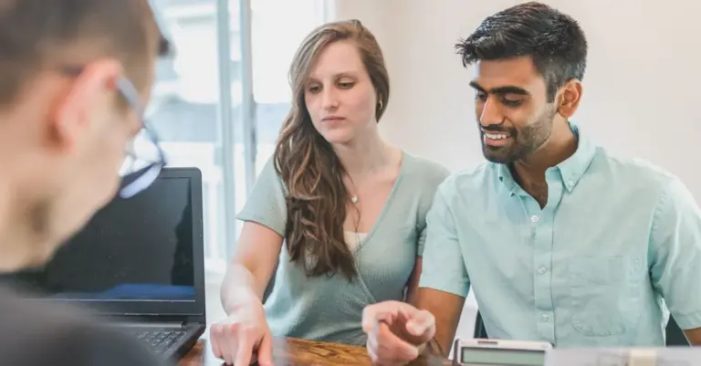Casal jovem a analisar documentos com calculadora ao lado de um consultor financeiro, comparando opções de crédito